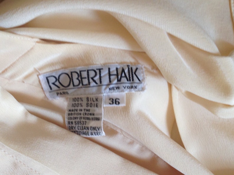 Op de afbeelding: Close-up van een cr&egrave;mekleurig zijden kledingstuk met een label met de tekst "ROBERT HAIK PARIS NEW YORK". Het label geeft aan dat het kledingstuk 100% zijde is, maat 36, en gemaakt in Hong Kong.