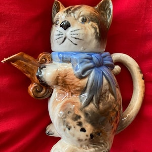 May include: A ceramic teapot shaped like a cat with brown, black and white fur. The cat is wearing a blue bow and has a brown spout.