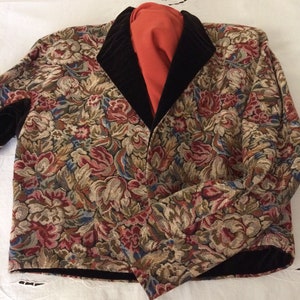 Op de afbeelding: Een jasje met bloemenpatroon met een zwarte fluwelen kraag en bies. De jas heeft een tapijtachtig ontwerp met rode, roze, blauwe en groene bloemen op een beige achtergrond. Onder de kraag is een oranje kledingstuk zichtbaar.