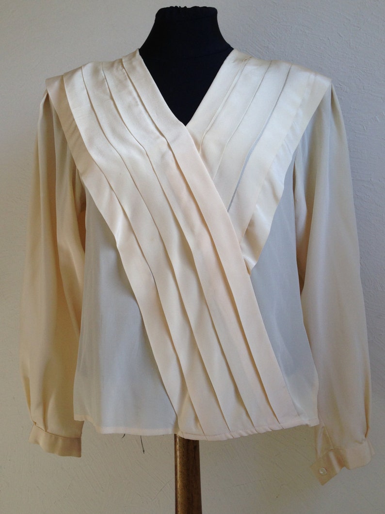 Op de afbeelding: Cr&egrave;mekleurige blouse met een V-hals. De blouse heeft lange mouwen en een geplooid, gedrapeerd accent over de voorkant. De stof lijkt een combinatie van satijn en chiffon te zijn, waardoor een gelaagd effect ontstaat. De blouse wordt gepresenteerd op een zwarte mannequin.