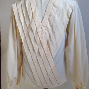 Op de afbeelding: Cr&egrave;mekleurige blouse met lange mouwen en een V-hals ontwerp. De achterkant heeft geplooide stoffen panelen die een gelaagd effect cre&euml;ren. De mouwen hebben manchetten met knopen en de blouse wordt op een mannequin getoond.