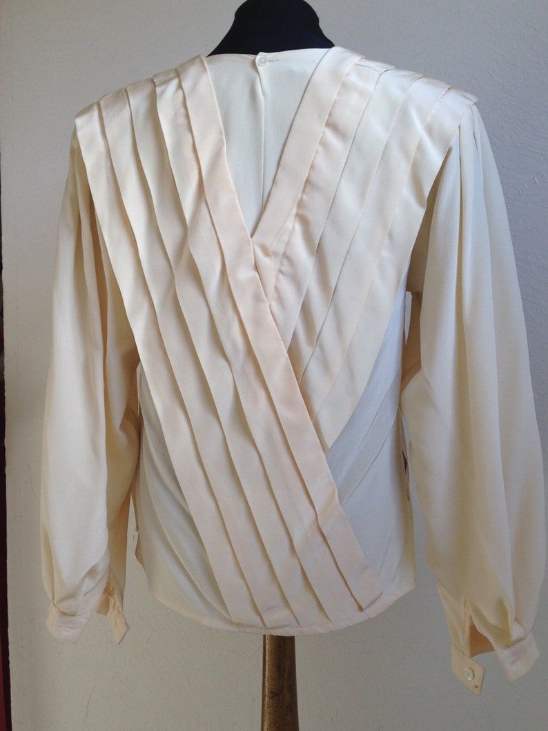 Op de afbeelding: Cr&egrave;mekleurige blouse met lange mouwen en een V-hals ontwerp. De achterkant heeft geplooide stoffen panelen die een gelaagd effect cre&euml;ren. De mouwen hebben manchetten met knopen en de blouse wordt op een mannequin getoond.