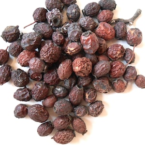May include: A collection of dried, dark reddish-brown berries with textured surfaces and small stems. The berries vary in size and shape, some with a slightly lighter hue. A few small twigs are also present, all against a white background.