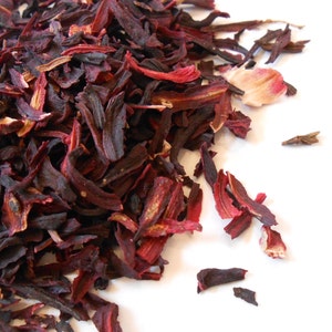 Può includere: Primo piano di fiori di ibisco essiccati, che mostrano tonalità rosso scuro e marrone. I petali e i sepali sono visibili, con alcuni pezzi più chiari sparsi tra quelli più scuri. L'immagine è su uno sfondo bianco, evidenziando la consistenza naturale.