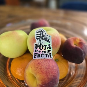 May include: A glass bowl filled with various fruits, including apples, oranges, and peaches. A sticker with the text "PUTA for your FRUTA" and a graphic of a hand squeezing a lemon is placed on top of an apple.
