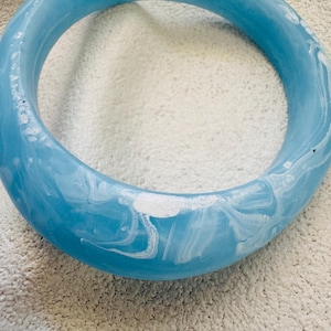 May include: A thick, circular bangle bracelet featuring a glossy finish with a marbled pattern of sky blue and white swirls. The jewellery piece has a smooth, rounded surface and a continuous, solid construction throughout its entire circumference.