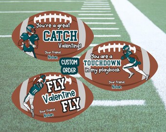 May include: Three brown and white football-shaped Valentine's Day cards with illustrations of football players wearing green and white uniforms. The text on the cards reads "You're a great CATCH Valentine! Your friend, Nolan", "You are a TOUCHDOWN in my playbook Your friend, Nolan", and "FLY Valentine FLY Your friend, Nolan".