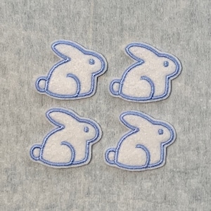 May include: Four white felt bunny rabbit patches with blue embroidered outlines. The patches are arranged in a 2x2 grid on a gray background.