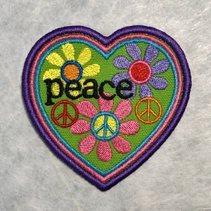 May include: A green heart-shaped patch with a peace sign and flowers embroidered on it. The word "peace" is embroidered in black letters.