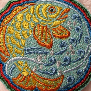 May include: A circular embroidered patch with a yellow and orange fish swimming in blue water. The patch is surrounded by a multi-colored border with red, orange, yellow, green, and blue.