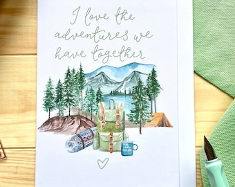 May include: A white card with a watercolor illustration of a forest scene with a lake, mountains, a backpack, a sleeping bag, a mug, and a tent. The text on the card reads "I love the adventures we have together."