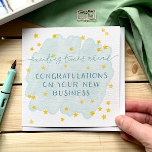 May include: A square greeting card with the text "exciting times ahead" and "Congratulations on your new business" in blue script. The card has a light blue watercolor background with yellow star accents. A light blue pen is visible on the left.