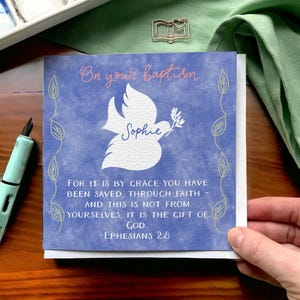 May include: A baptism greeting card with a blue watercolour background. The card features a white dove holding an olive branch, the name "Sophie", and the text "On your baptism". The card also includes a Bible verse.