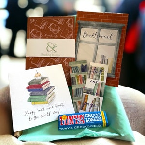 May include: A gift basket for book lovers with a brown and white reading journal, a brown and white booktrovert journal, a bookmark with a stack of books, a card with a stack of books and the text "Happy 'add more books to the shelf' day!" and a blue chocolate bar with the text "TONY'S CHOCOLONELY."