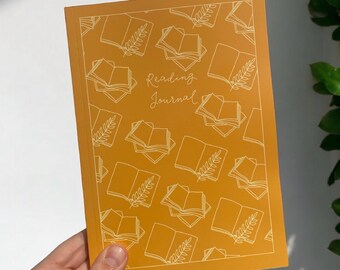 May include: A mustard yellow reading journal with a white outline of stacked books and a leaf design. The text "Reading Journal" is written in white cursive script. The journal is designed to fit 100 books.