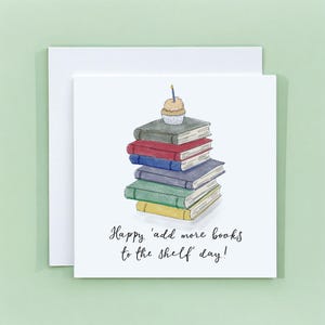 May include: A square greeting card featuring a watercolour illustration of a stack of five books in grey, red, blue, purple, and green, topped with a cupcake with a single blue candle. The text reads, Happy 'add more books to the shelf' day!