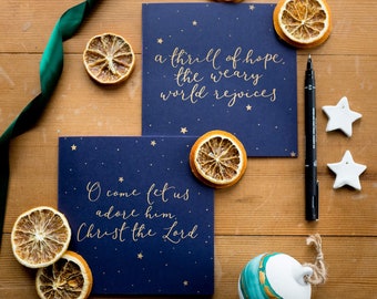 May include: Two blue Christmas cards with gold foil stars and text. The top card reads "a thrill of hope, the weary world rejoices". The bottom card reads "O come let us adore him, Christ the Lord".