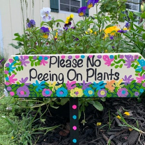 May include: A white wooden sign with a colorful floral border and the text "Please No Peeing On Plants". The sign is attached to a black stake with pink, blue, and black polka dots.