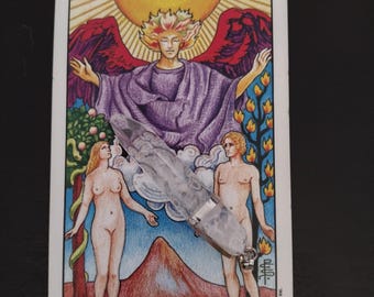 May include: A tarot card depicting "The Lovers" with a central figure, angel with red wings, and two nude figures. A clear crystal wand is placed over the card. The card has a white border and the text "THE LOVERS."