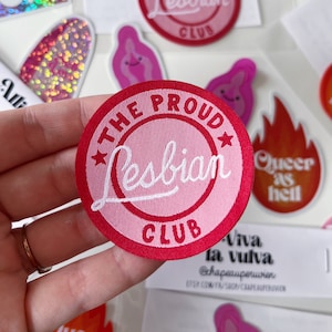 Op de afbeelding: Een ronde, geborduurde patch in roze en rood met de tekst "The Proud Lesbian Club" in witte letters. De patch is rond en heeft aan beide kanten van de tekst een witte ster.
