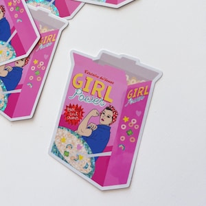 Peut inclure: Autocollant rose représentant une boîte de céréales avec les mots "GIRL POWER" en jaune et bleu. L'illustration montre une femme musclée avec le texte "Pour tout cramer". D'autres autocollants similaires sont visibles.