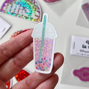 Peut inclure: Un autocollant holographique représentant un bubble tea rose avec des perles de boba colorées et une paille verte. L'autocollant est sur un fond blanc.