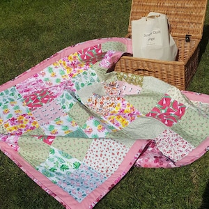 May include: A pink and green patchwork quilt with a variety of floral and geometric patterns. The quilt is folded and lying on a green grassy surface. A wicker picnic basket with a white tote bag that says "Scrappy Quilt Range" is in the background.