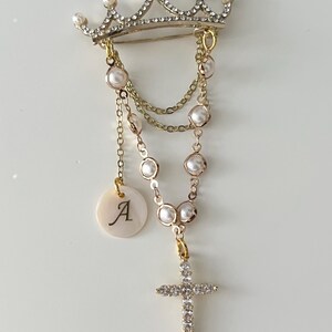 May include: A gold-tone crown brooch featuring clear rhinestones and white faux pearls. Dangling from the crown are gold chains adorned with faux pearls, a circular white charm with the letter A, and a cross pendant encrusted with clear rhinestones.