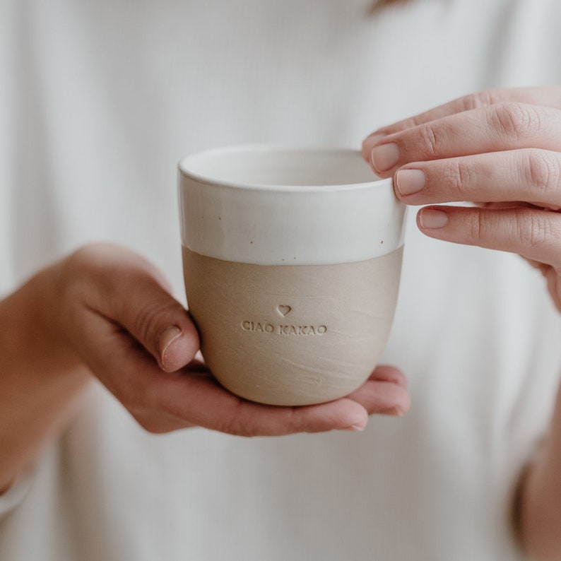 May include: A ceramic mug with a white interior and a beige exterior. The mug has a heart and the text 'CIAO KAKAO' printed on the exterior.