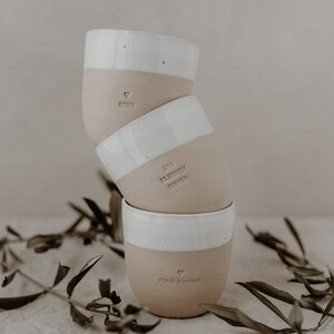 May include: Three stacked ceramic mugs with white rims and a light brown body. The top mug has the word 'MINI' and a heart symbol. The middle mug has the words 'DIE KLEINEN DINGE'. The bottom mug has the words 'CIAO KAYO' and a heart symbol.