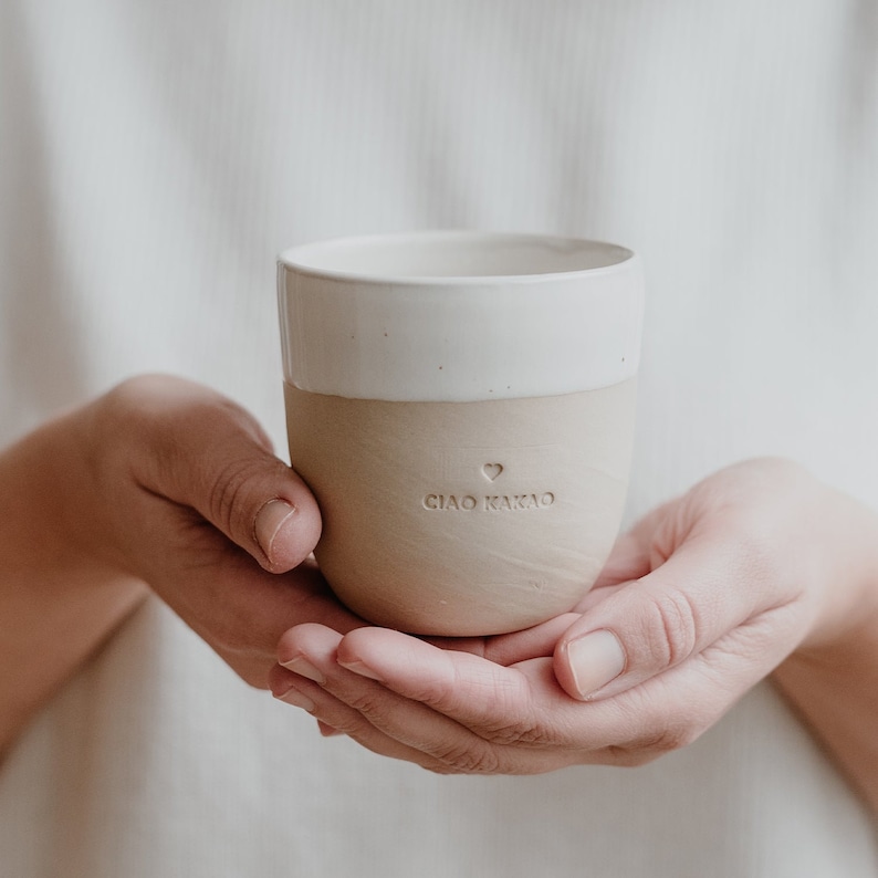 May include: A ceramic mug with a white top and a beige bottom. The mug has a heart and the text 'CIAO KAKAO' printed on the bottom.