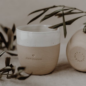 May include: A white and beige ceramic mug with the words 'Ciao Kakao' and a heart symbol on the side. The mug is sitting on a beige surface with a green plant branch behind it.