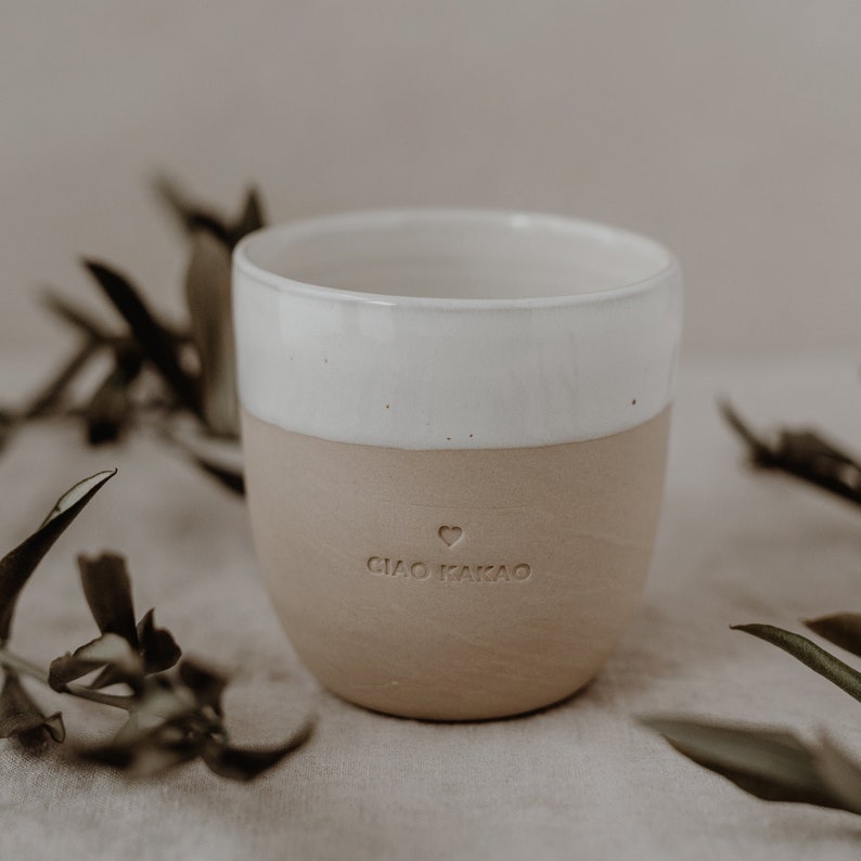 May include: A white and beige ceramic mug with the words 'Ciao Kakao' and a heart symbol on the bottom.