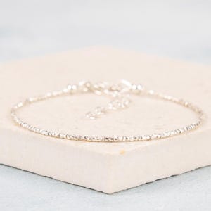 May include: A delicate silver bracelet with small, square beads. The bracelet is displayed on a light beige surface against a soft blue background. The clasp is visible, suggesting an adjustable fit.