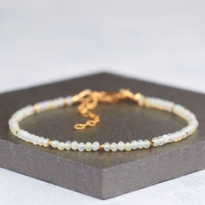 May include: A fine opal bead bracelet with a gold-coloured chain and clasp. The bracelet is made of small, round, iridescent opal beads. The gold chain and clasp add a touch of elegance. The bracelet is displayed on a dark grey surface.