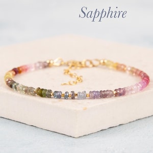 Op de afbeelding: Een delicate armband met een streng van kleurrijke saffier edelstenen. De armband heeft een goudkleurige ketting en sluiting. De edelstenen zijn in tinten roze, paars, groen en geel. Het woord "Sapphire" is cursief geschreven.