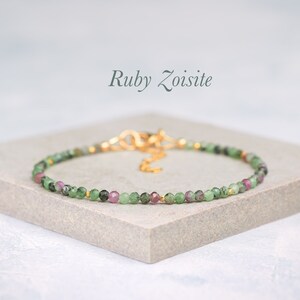 May include: A delicate bracelet made with small green and pink ruby zoisite beads and gold accents. The bracelet is on a grey surface.