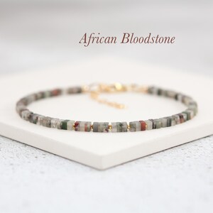 May include: A delicate bracelet featuring small, square-shaped African Bloodstone beads. The beads are a mix of green, grey, and red, with gold accents. The bracelet has an adjustable clasp.