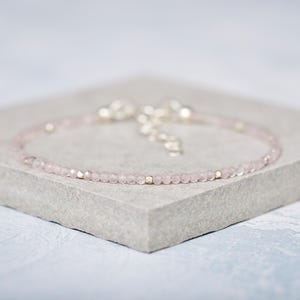 May include: A delicate rose quartz bracelet, featuring small, faceted beads and silver-toned details. The bracelet is displayed on a grey, textured surface. A beautiful piece of jewellery.