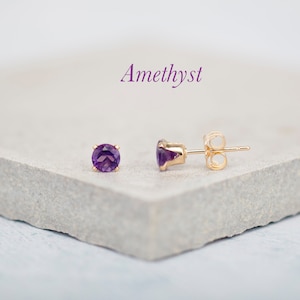 Könnte beinhalten: Ein Paar goldene Ohrstecker mit runden Amethyst-Edelsteinen. Die Ohrringe liegen auf einer hellgrauen Oberfläche. Amethyst.