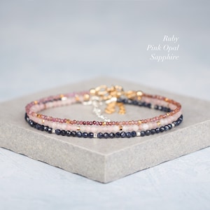Puede incluir: Un conjunto de tres pulseras de cuentas delicadas con piedras preciosas facetadas de rub&iacute;, &oacute;palo rosa y zafiro. Las pulseras est&aacute;n acentuadas con peque&ntilde;as cuentas met&aacute;licas e incluyen un cierre de metal. El texto dice: Ruby, Pink Opal, Sapphire.