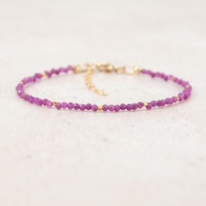 May include: A delicate bracelet featuring a continuous strand of small, faceted, deep magenta-coloured gemstone beads. The design is accented with tiny, polished gold-toned metal spacers at regular intervals and includes a gold-toned metal chain extender.