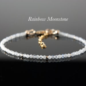 May include: A delicate bracelet featuring small, clear beads strung together. The bracelet has a gold-colored clasp and extender chain. The text "Rainbow Moonstone" is displayed in white script above the bracelet.