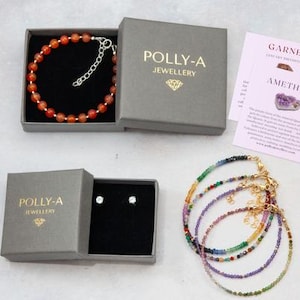 Puede incluir: Una colecci&oacute;n de joyer&iacute;a que incluye una pulsera de cuentas naranjas en una caja gris, un par de pendientes en una caja gris y cuatro pulseras de cuentas multicolores. Las cajas tienen la etiqueta POLLY-A JEWELLERY con un icono de diamante.