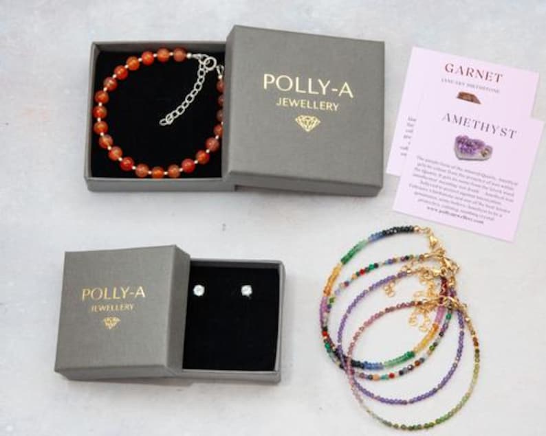 Puede incluir: Una colecci&oacute;n de joyer&iacute;a que incluye una pulsera de cuentas naranjas en una caja gris, un par de pendientes en una caja gris y cuatro pulseras de cuentas multicolores. Las cajas tienen la etiqueta POLLY-A JEWELLERY con un icono de diamante.