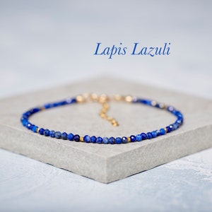 Może przedstawiać: Delikatny bransoletka wykonana z małych, ciemnoniebieskich koralików lapis lazuli i złotych akcentów. Bransoletka leży na szarej powierzchni. "Lapis Lazuli" jest napisane niebieskim kolorem nad bransoletką.