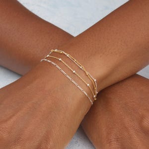 May include: Three delicate bracelets in gold and silver tones are stacked on a wrist. The bracelets feature different chain styles, including a beaded chain, a figaro chain, and a delicate chain.