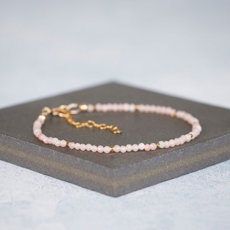 Puede incluir: Una pulsera delicada con peque&ntilde;as cuentas facetadas de color rosa p&aacute;lido dispuestas en un patr&oacute;n circular continuo. El dise&ntilde;o est&aacute; acentuado con diminutos espaciadores de metal dorado reflectante e incluye una cadena extensora dorada.