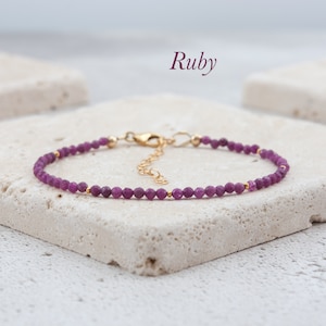May include: A delicate gold chain bracelet with small, faceted ruby beads. The bracelet is on a white background and the word "Ruby" is written above it.