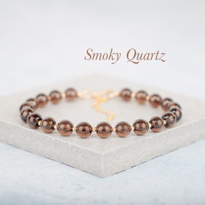 May include: A bracelet made of smoky quartz beads, each a dark brown colour. The bracelet has gold-coloured details and a fine chain. The text "Smoky Quartz" is written in a stylish font.